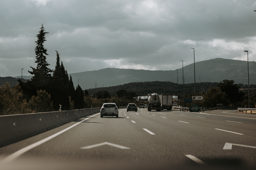 Cars travel on a highway through a scenic mountain landscape under cloudy skies.