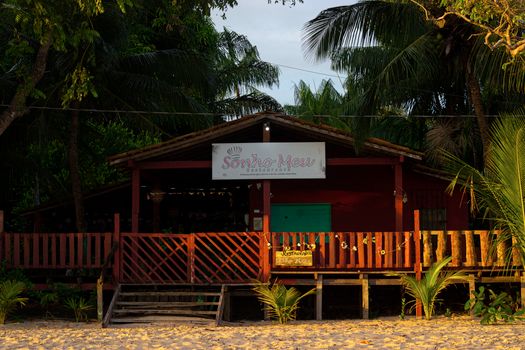Photo by Marlon Costa Charming beachfront restaurant surrounded by palm trees at sunset, with sandy beach view.