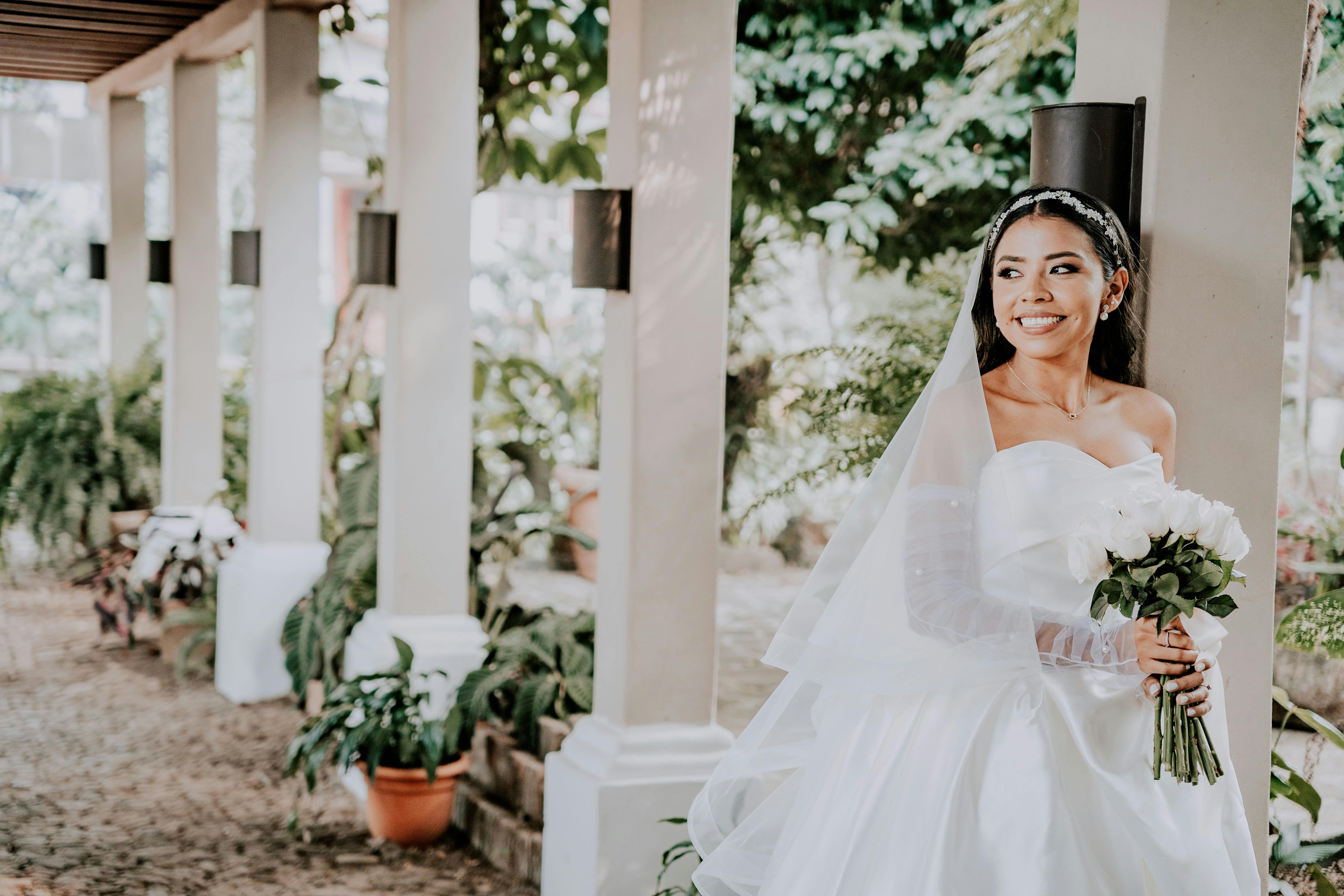 Smiling bride in a white dress holding a bouquet, posing in a garden colonnade.
