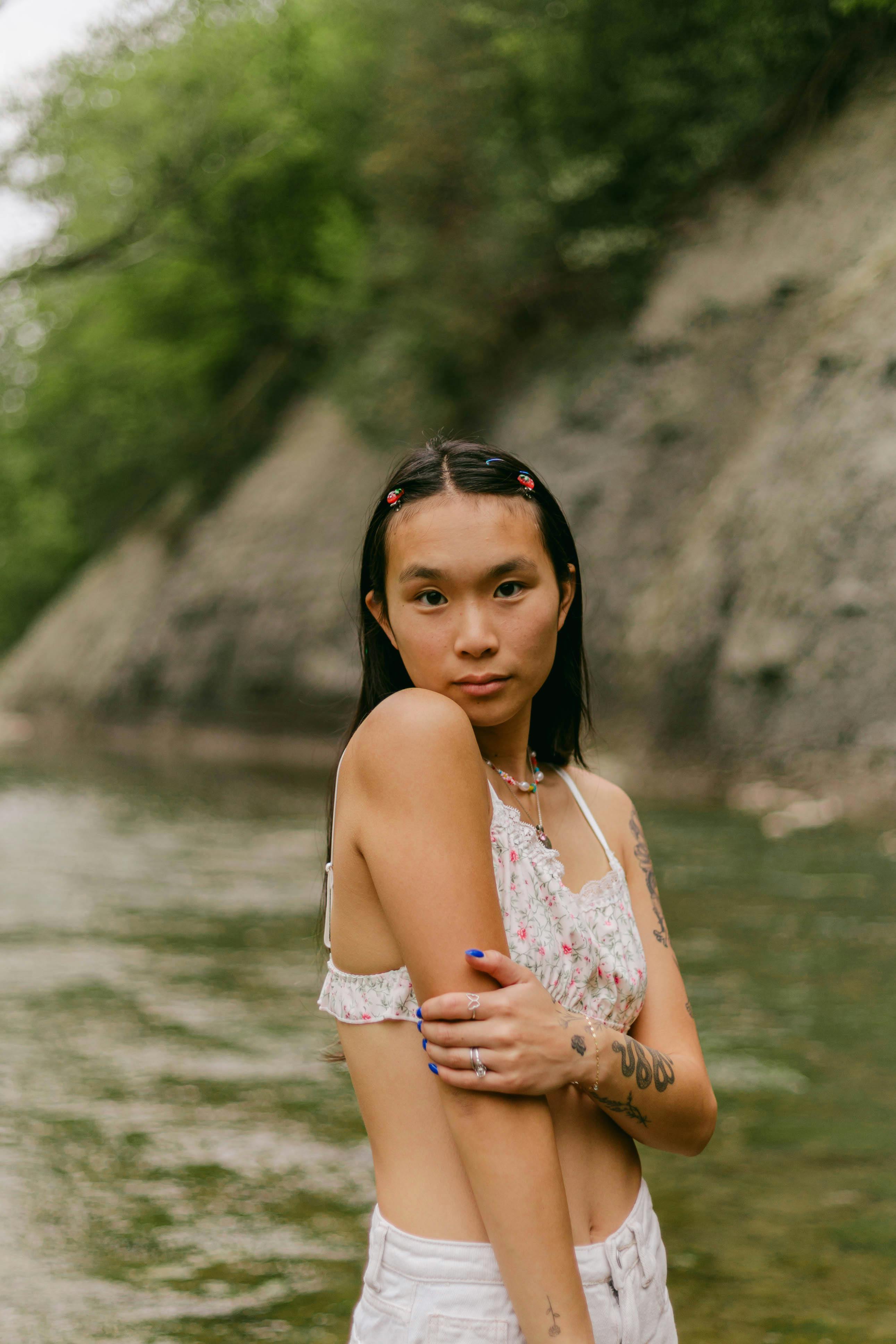 A young woman with tattoos poses confidently by a peaceful river in summer.