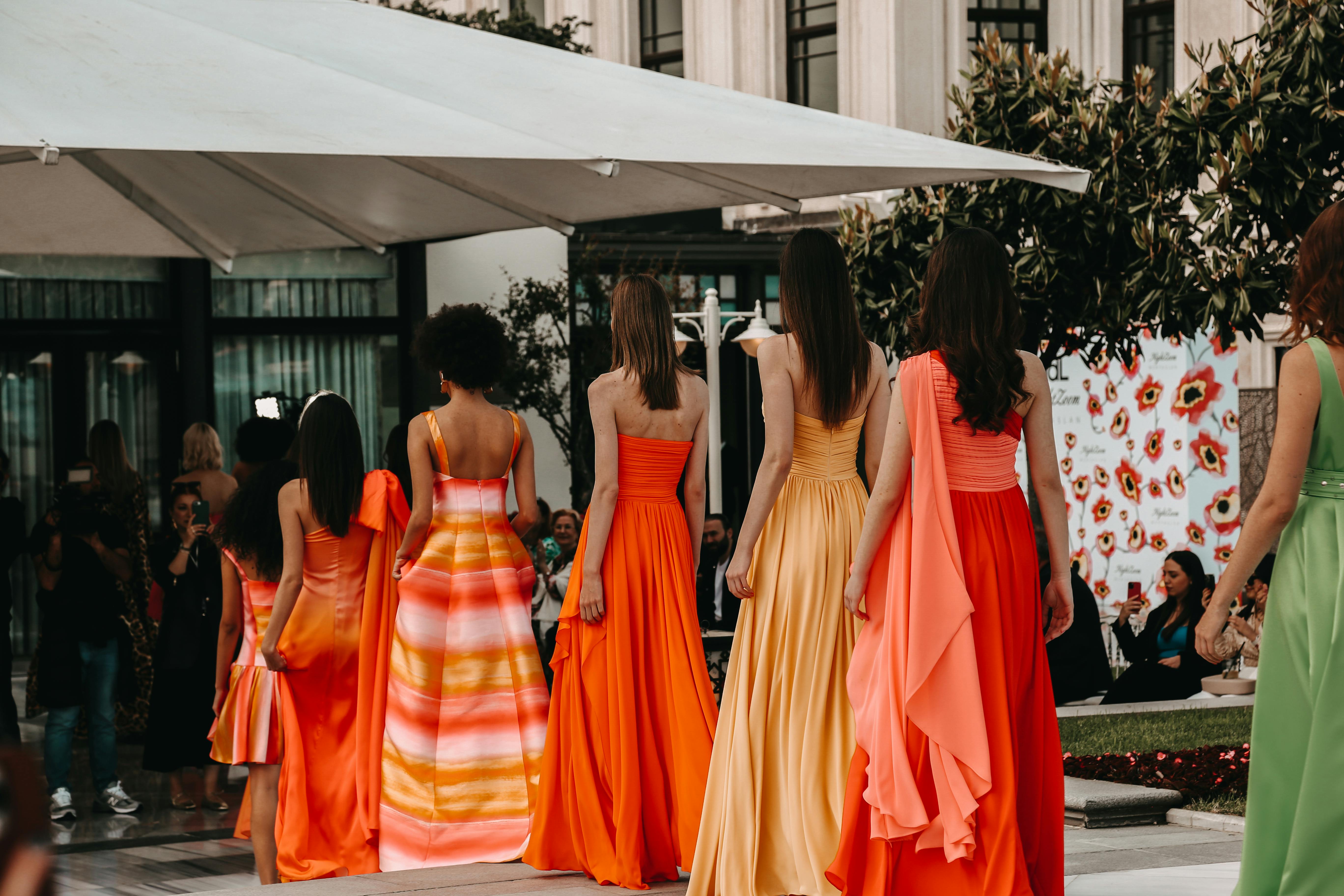 Models in stunning vibrant dresses walk the runway in an outdoor fashion show.
