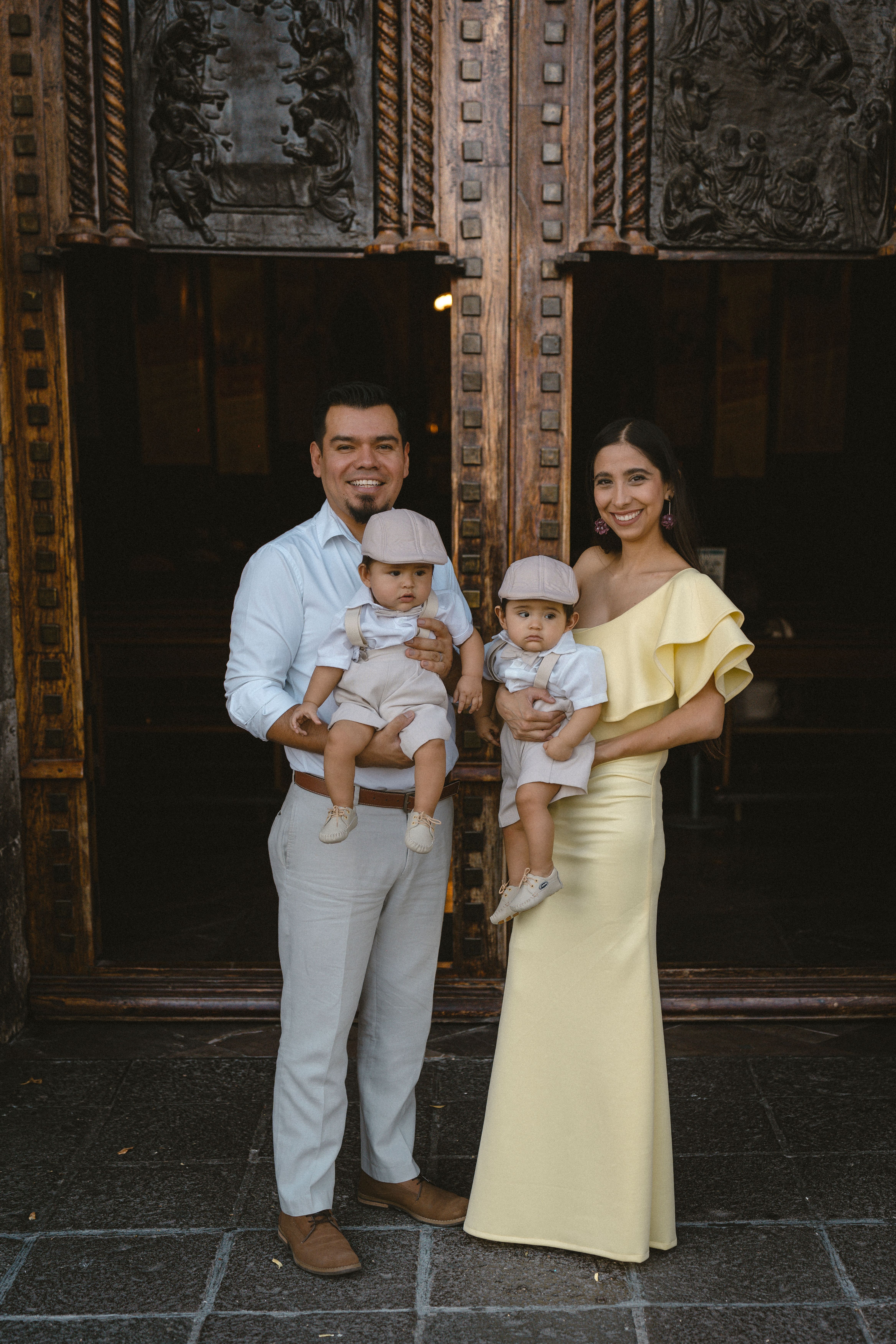 A couple standing outside a church, holding twin babies, captured in a family portrait.