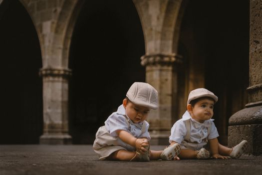 Charming portrait of twin babies in matching vintage outfits sitting on a stone floor against an archway background.