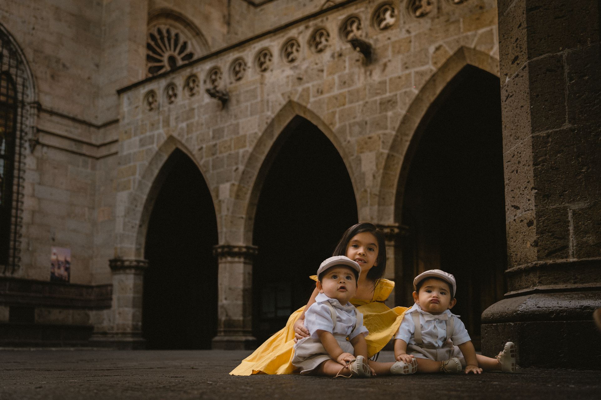 Baby Twins in Matching Outfits and Their Sister Sitting in a Castle Courtyard
