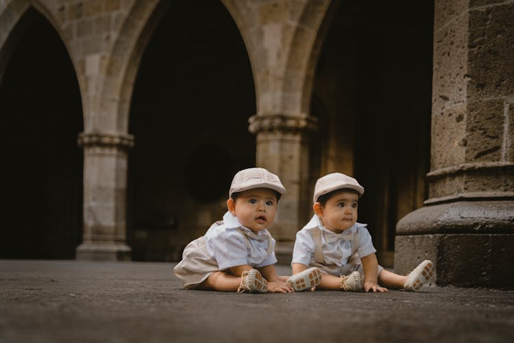 Baby Twins In Matching Outfits Sitting In A Castle Courtyard 