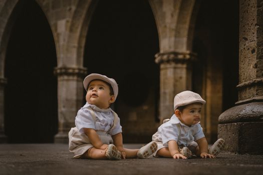 Adorable twin babies in vintage attire sitting under a rustic stone archway, capturing innocence and charm.