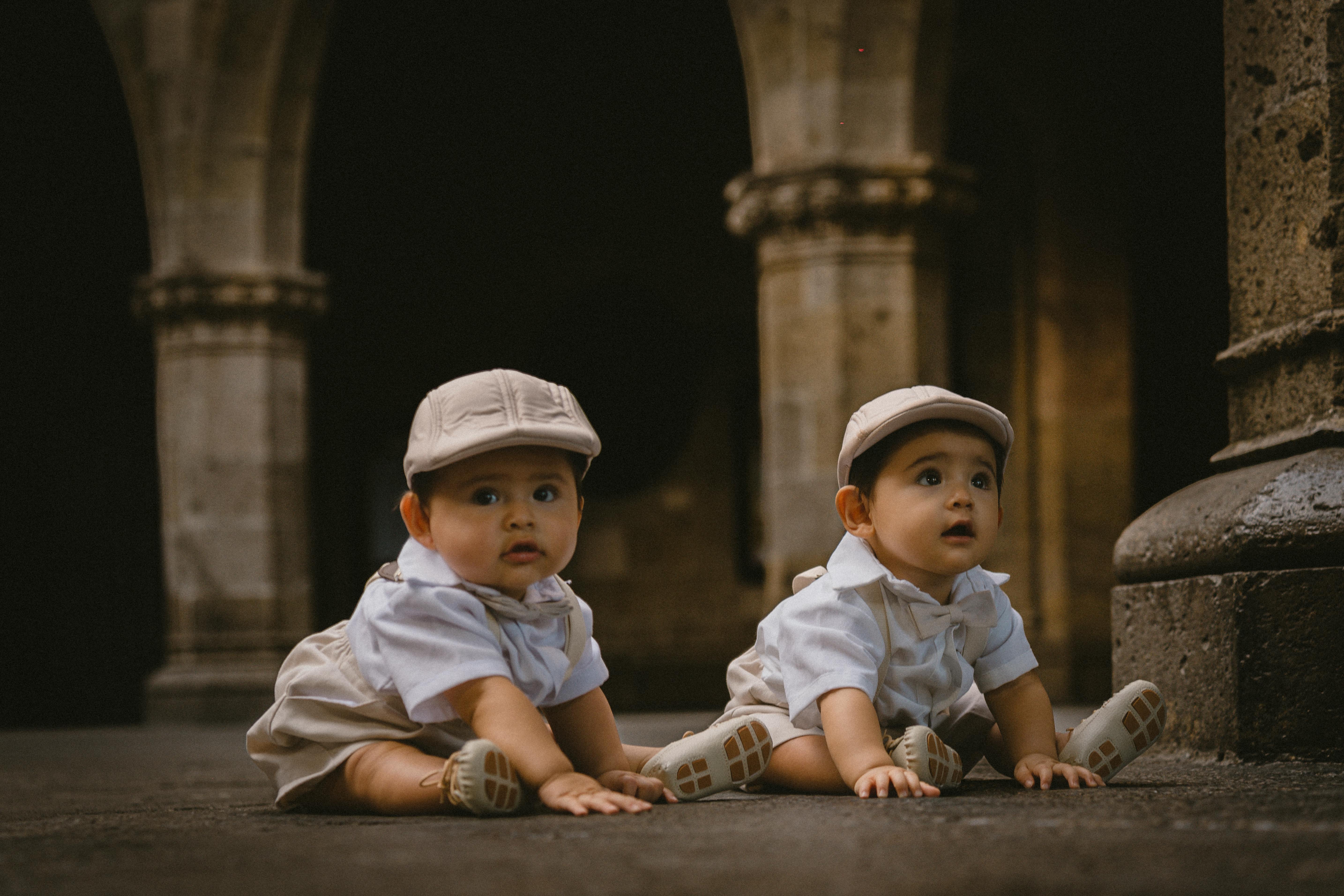 Adorable twin babies in caps sitting on an ancient stone pavement with arches in the background.