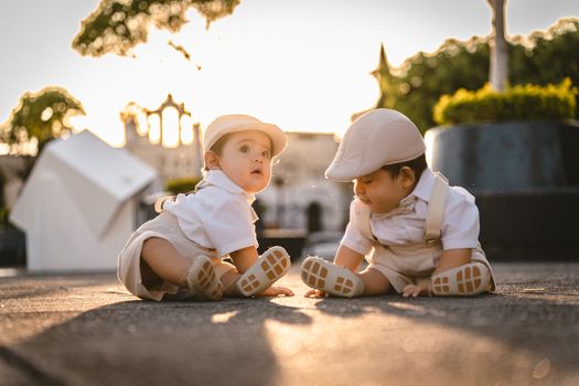 Cute twin babies in white outfits sitting outdoors at sunset, exuding warmth.