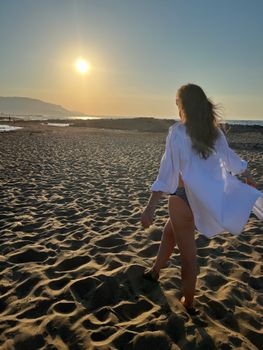A woman in a white shirt walks along a sandy beach at sunset, embracing a tranquil vacation moment.