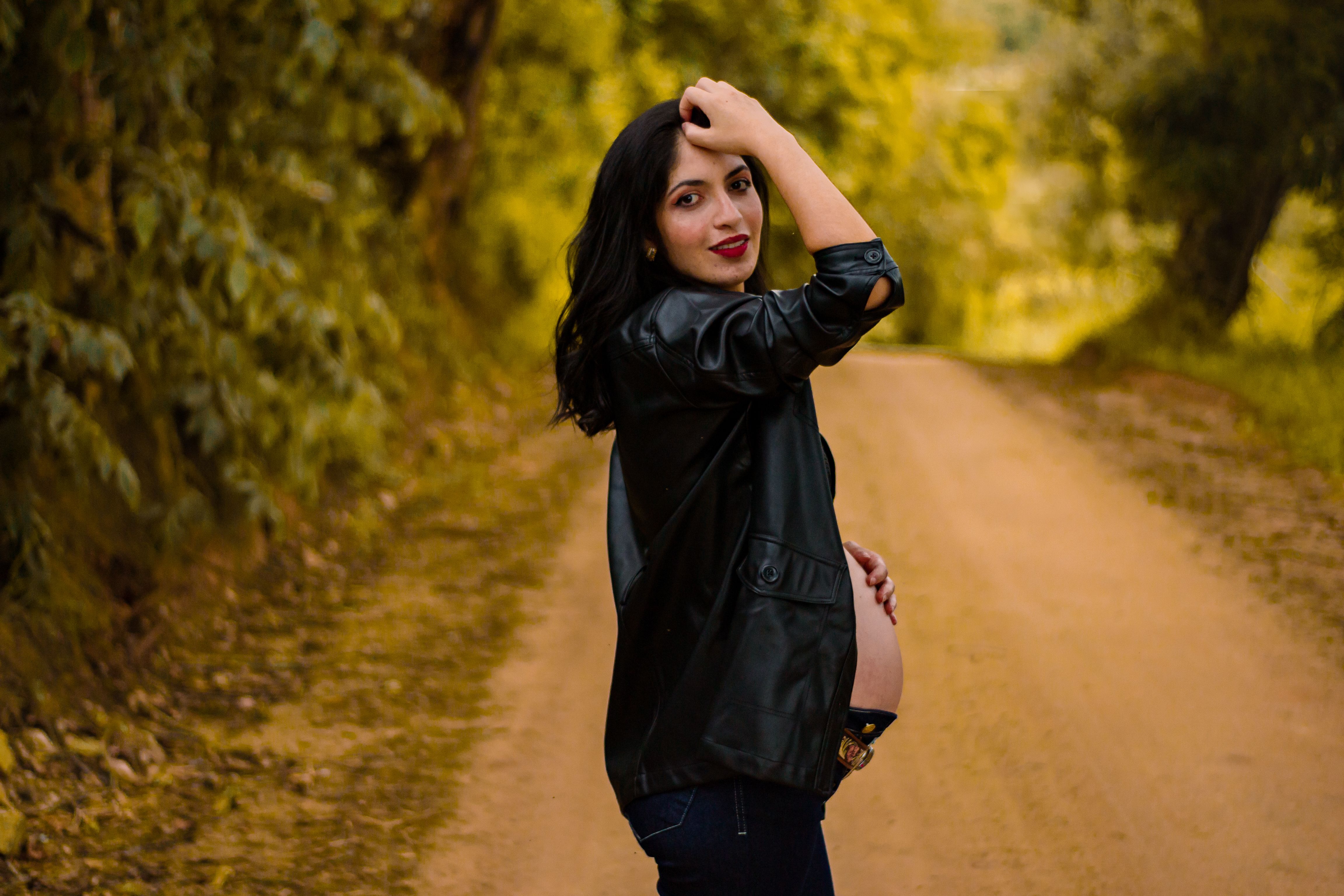 Caucasian pregnant woman in a black jacket posing on a dirt road surrounded by nature.