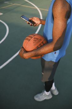 Athlete holding a basketball and smartphone on an outdoor court during daytime.