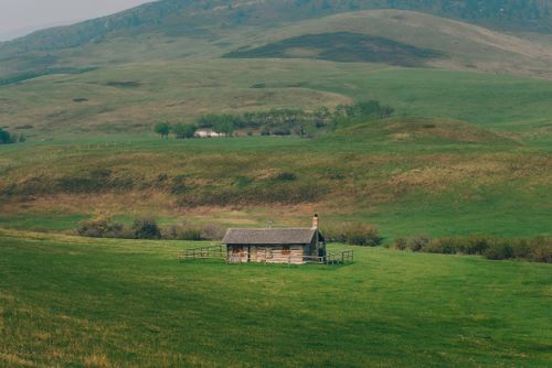 Free A charming wooden cabin nestled amidst expansive green hills and fields in a serene rural setting. Stock Photo