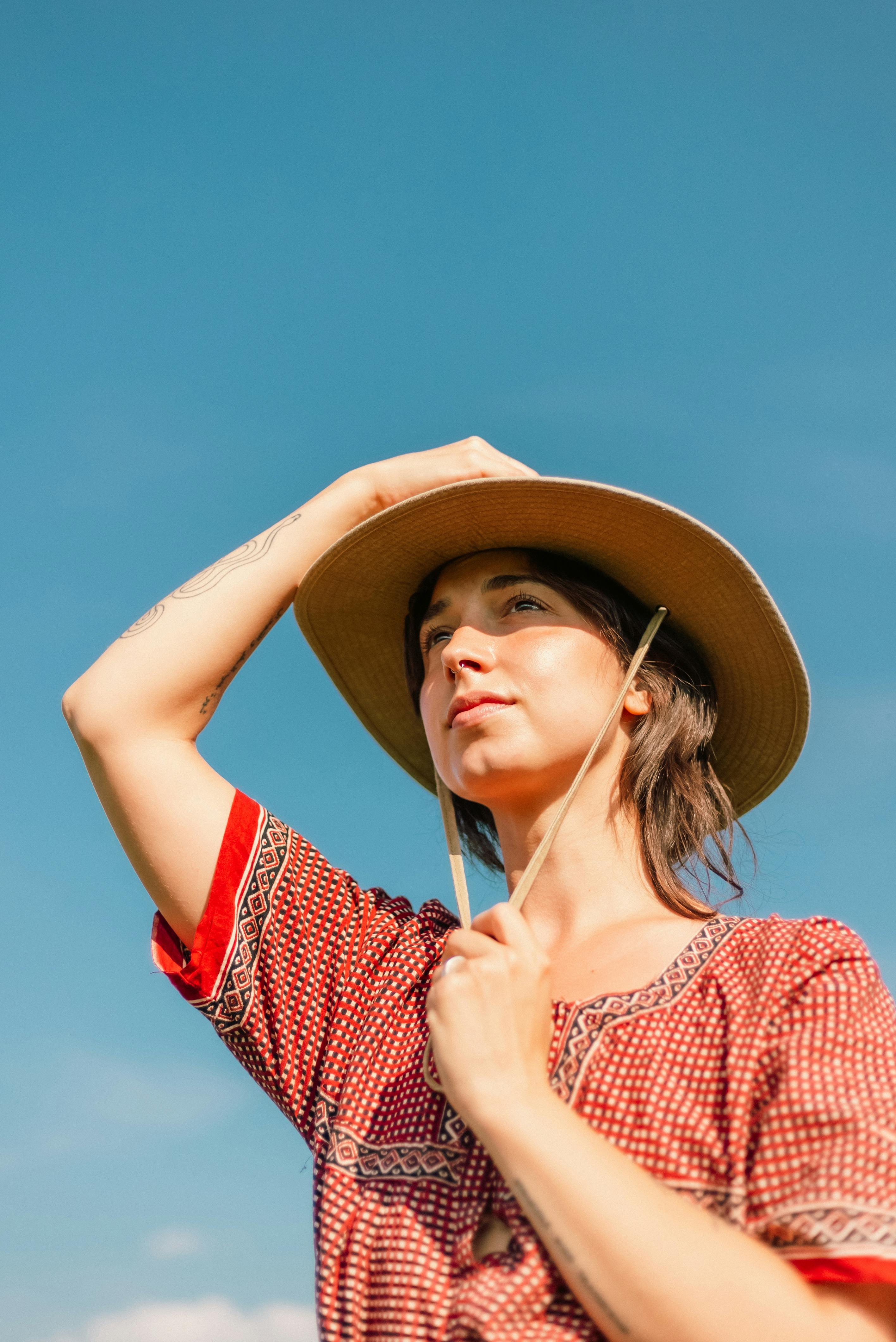 Young woman wearing a hat in a patterned dress gazing at the clear blue sky on a sunny day.
