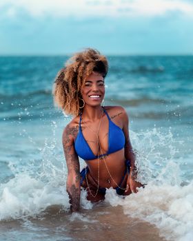 Joyful woman with curly hair in blue bikini enjoying the beach waves on a sunny day.