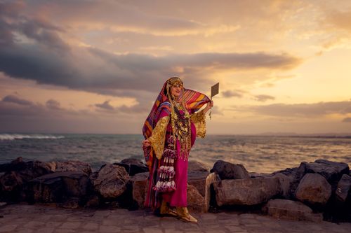 Free A woman in traditional Tunisian attire poses on Hammamet shore against a striking sunset background. Stock Photo