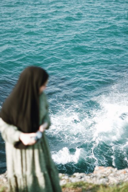 A woman in a hijab stands near the edge of a cliff overlooking the ocean waves below.