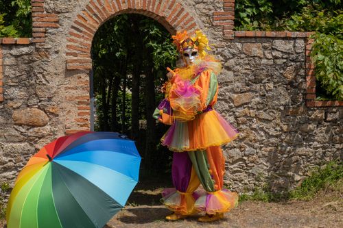 Free A colorful jester props with a rainbow umbrella by a stone arch in Clisson, France. Stock Photo