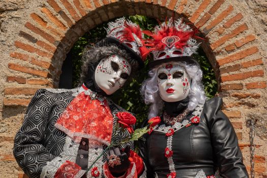 Two people in elaborate Venetian-style costumes and masks posing by a stone archway in Clisson, France.