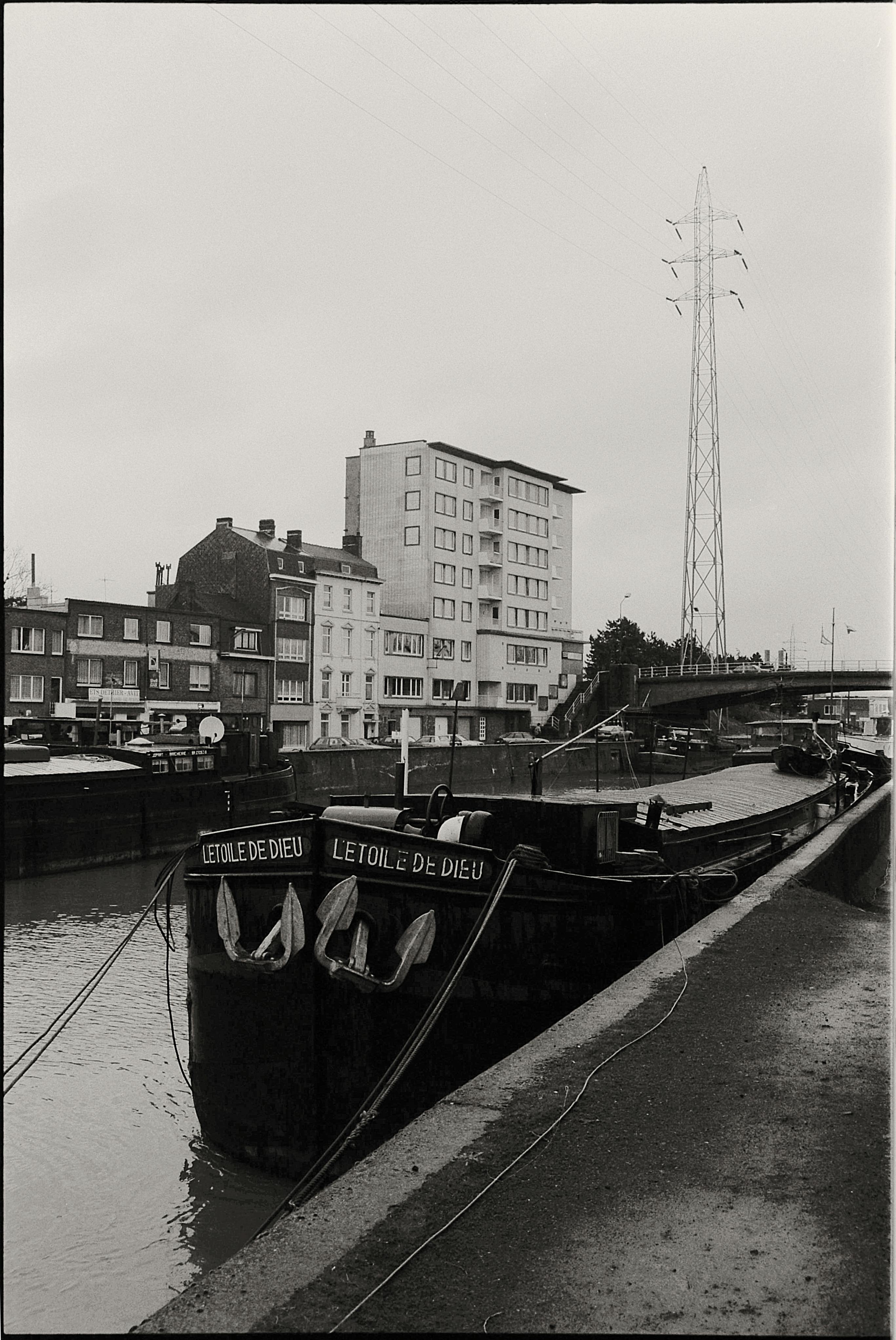 Black and white photo of a barge moored along an urban canal with buildings.