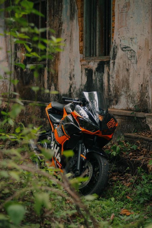 Free A vibrant orange motorbike parked beside an old, weathered building wall with green plants. Stock Photo