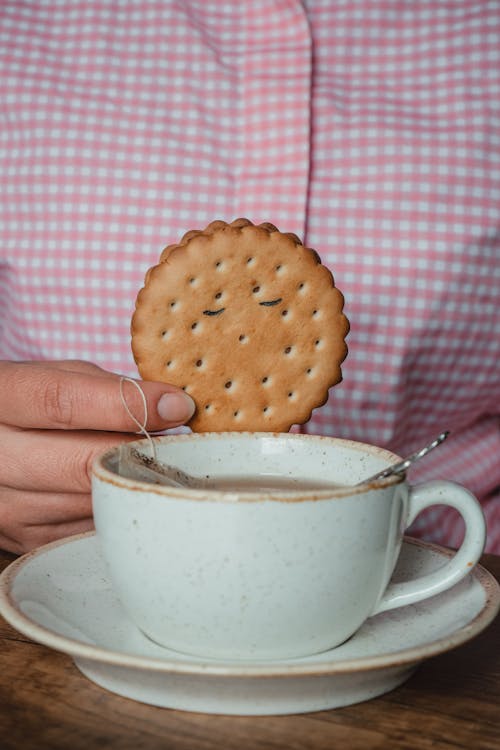 Person Holding a Cookie by Porcelain Cup of Tea