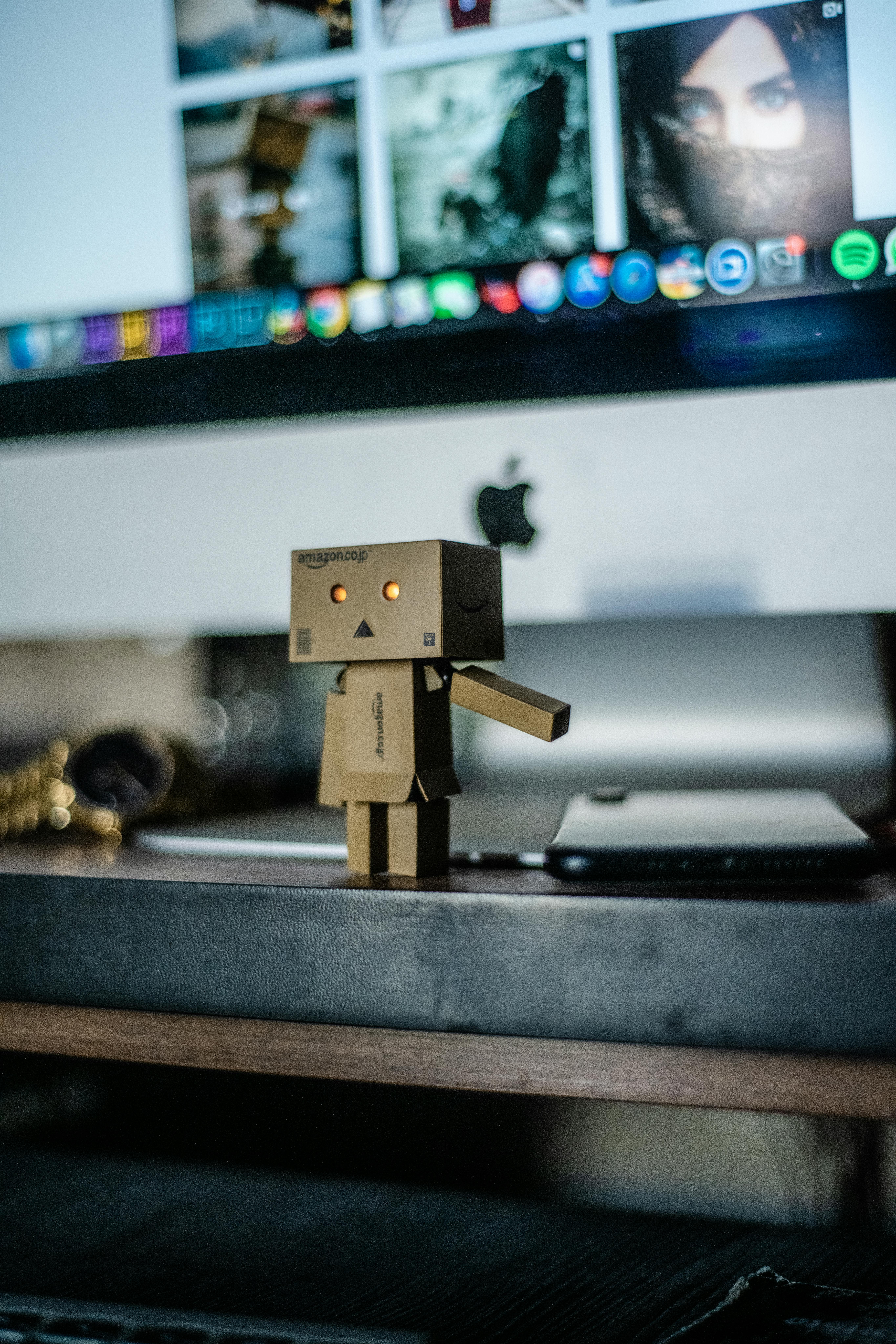 Cardboard robot toy stands on desk with computer screen backdrop, creating a tech and play ambiance.