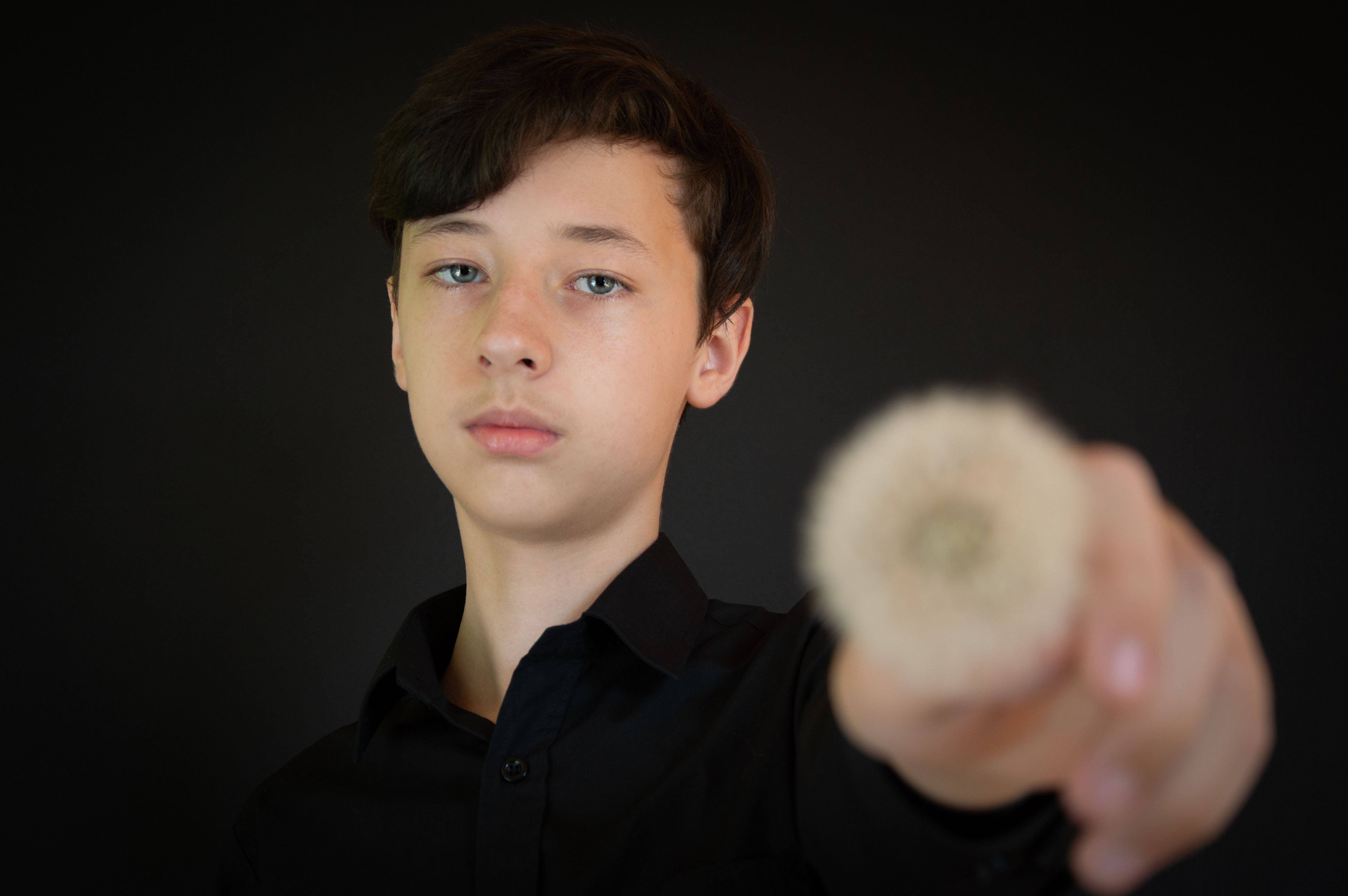 Teenage boy holds a dandelion against a black background, offering a contemplative expression.