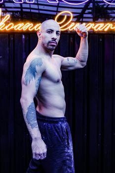 A muscular man with tattoos confidently poses against a neon-lit background, showcasing strength and style.