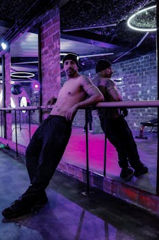 Muscular man with tattoos in a modern industrial gym leaning on a barre with violet lighting.