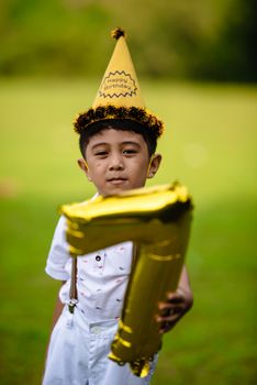 A young boy celebrating his birthday outdoors with a gold number one balloon and party hat.