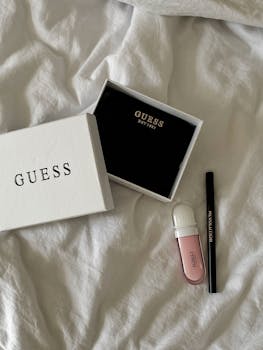 Top view of Guess accessories box with makeup essentials on white fabric. Chic and modern aesthetic.