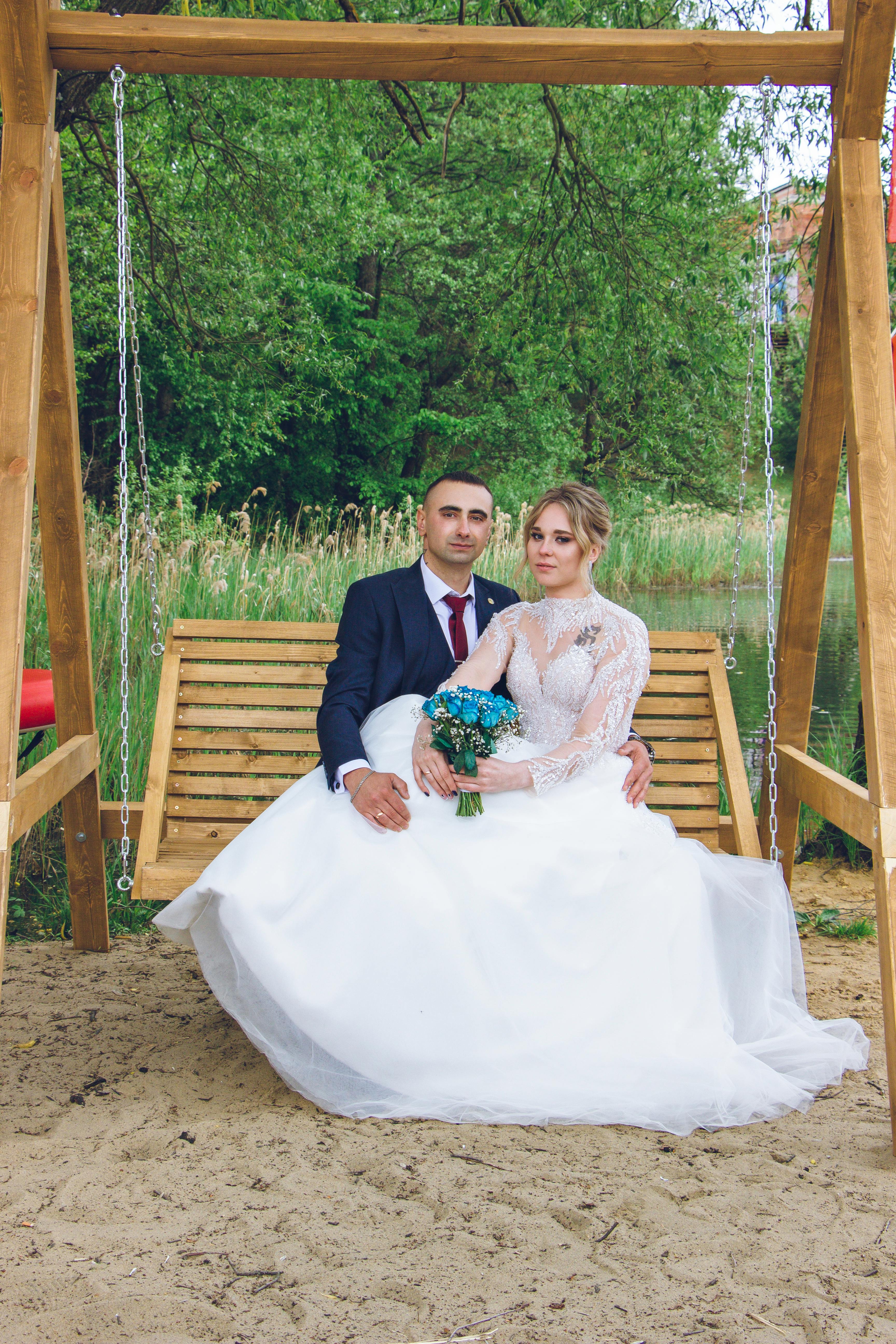 Newlyweds sit together on a wooden swing in a serene outdoor setting, exuding elegance and love.