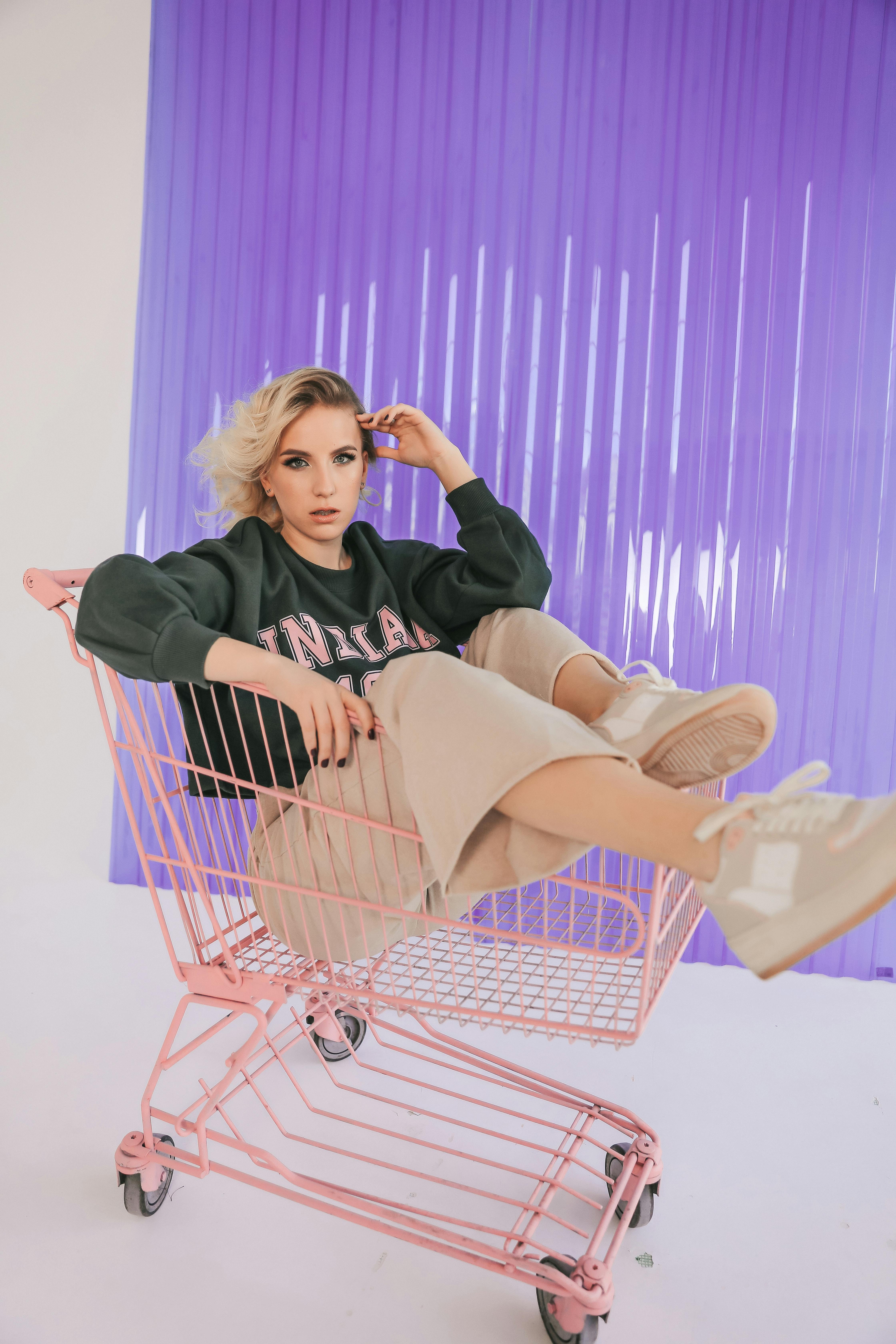 Trendy woman relaxing in a pink shopping cart with a purple backdrop.