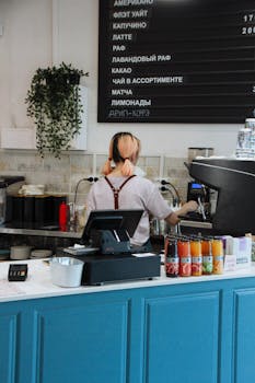 A barista making coffee behind a stylish counter in a modern cafe with a variety of drinks displayed.