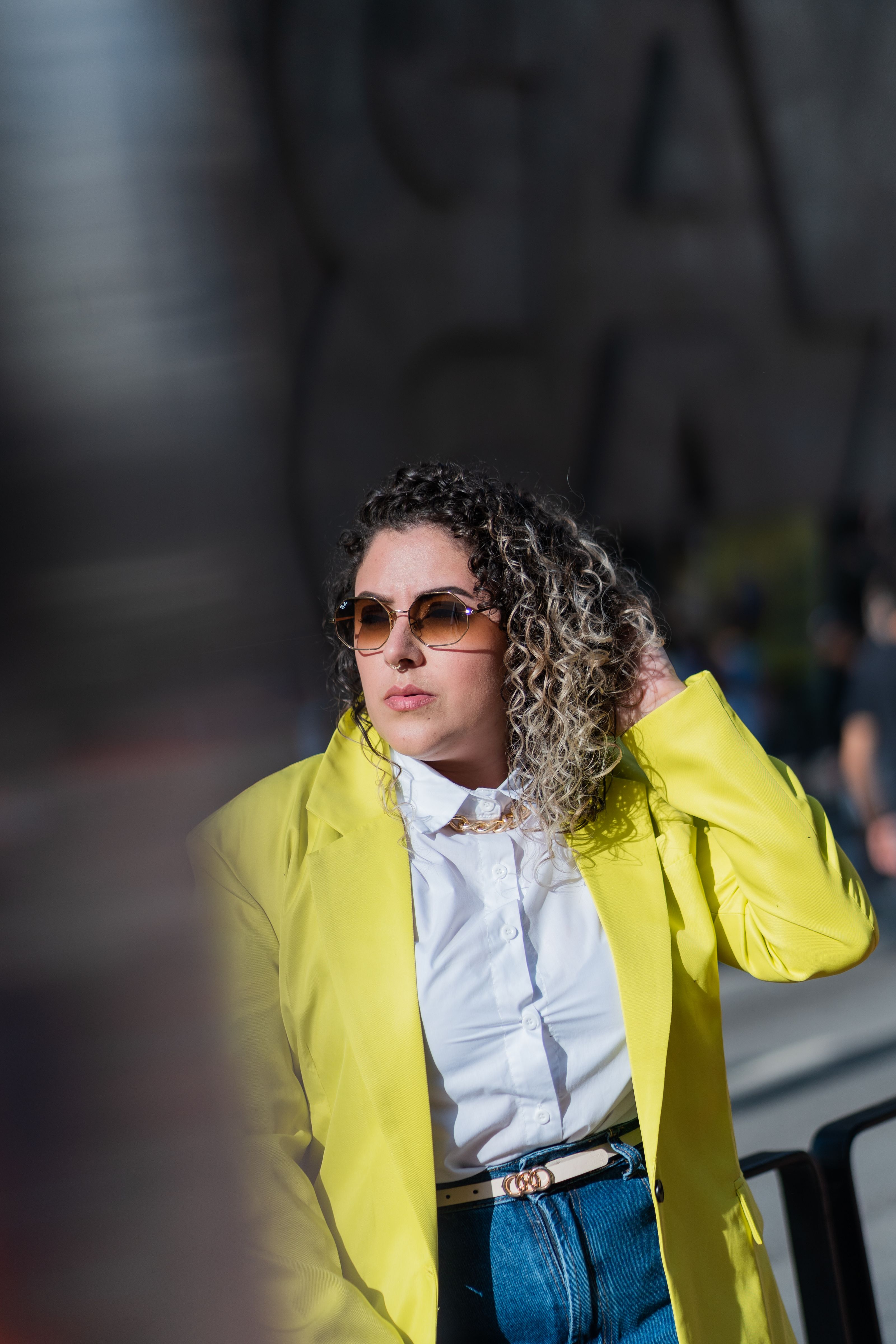 Mulher estilosa usando óculos de sol, casaco amarelo e jeans denim ao ar livre durante o dia.