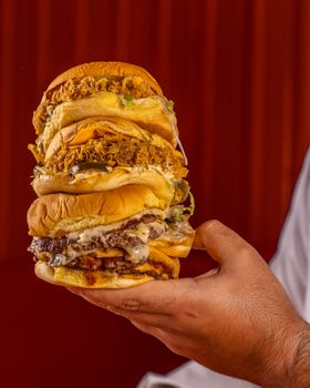 Close-up of a hand holding a stack of gourmet burgers with various toppings against a red background.