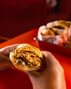Close-up of a person holding a delicious fried chicken sandwich on a red table, with a tray of similar items in the background.