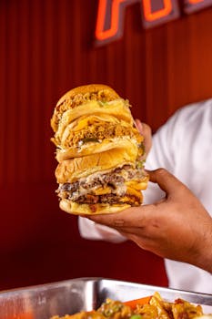 A person holding multiple stacked burgers in a lively restaurant atmosphere.