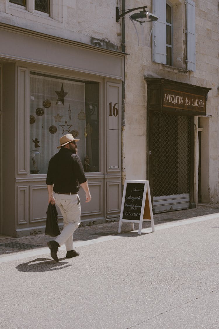 Man Wearing A Summer Hat Walking On A Street By A Cafe With Chalkboard Sign