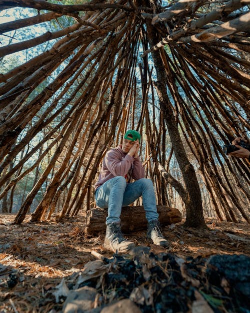 Man in a Pink Hoodie and Jeans Sitting on a Log in a Forest Shelter