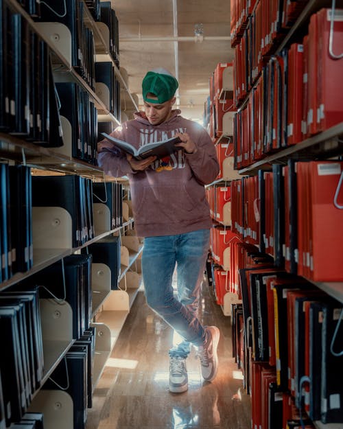 Man in a Pink Hoodie and Jeans Reading a Book Between the Bookcases in the Library