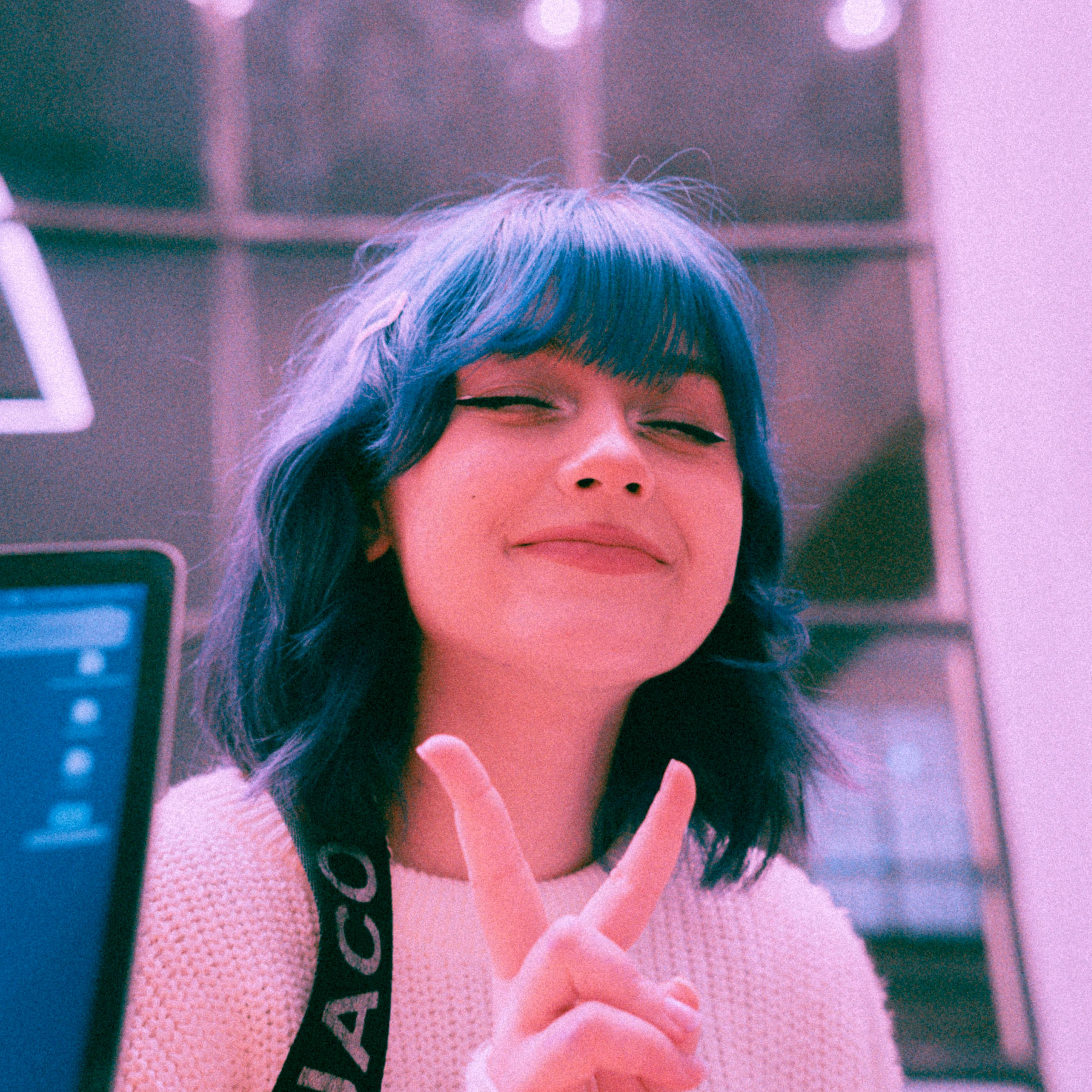 Happy teenager with vibrant blue hair smiling and flashing peace sign indoors.