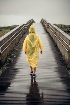 A person in a yellow raincoat walks on a wet boardwalk on a rainy day.