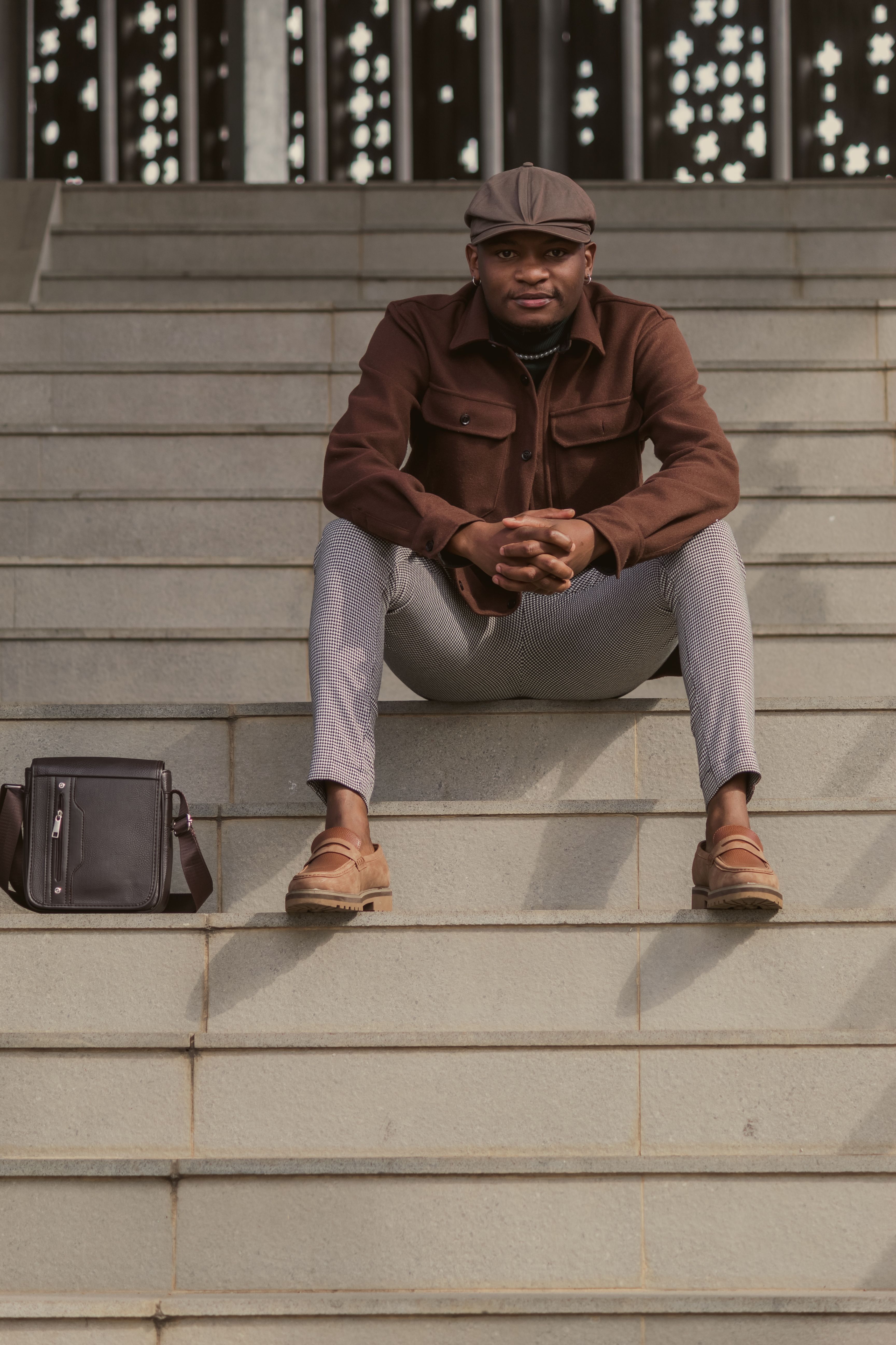 A fashionable man in a brown jacket and cap sitting on steps with a black bag beside him.