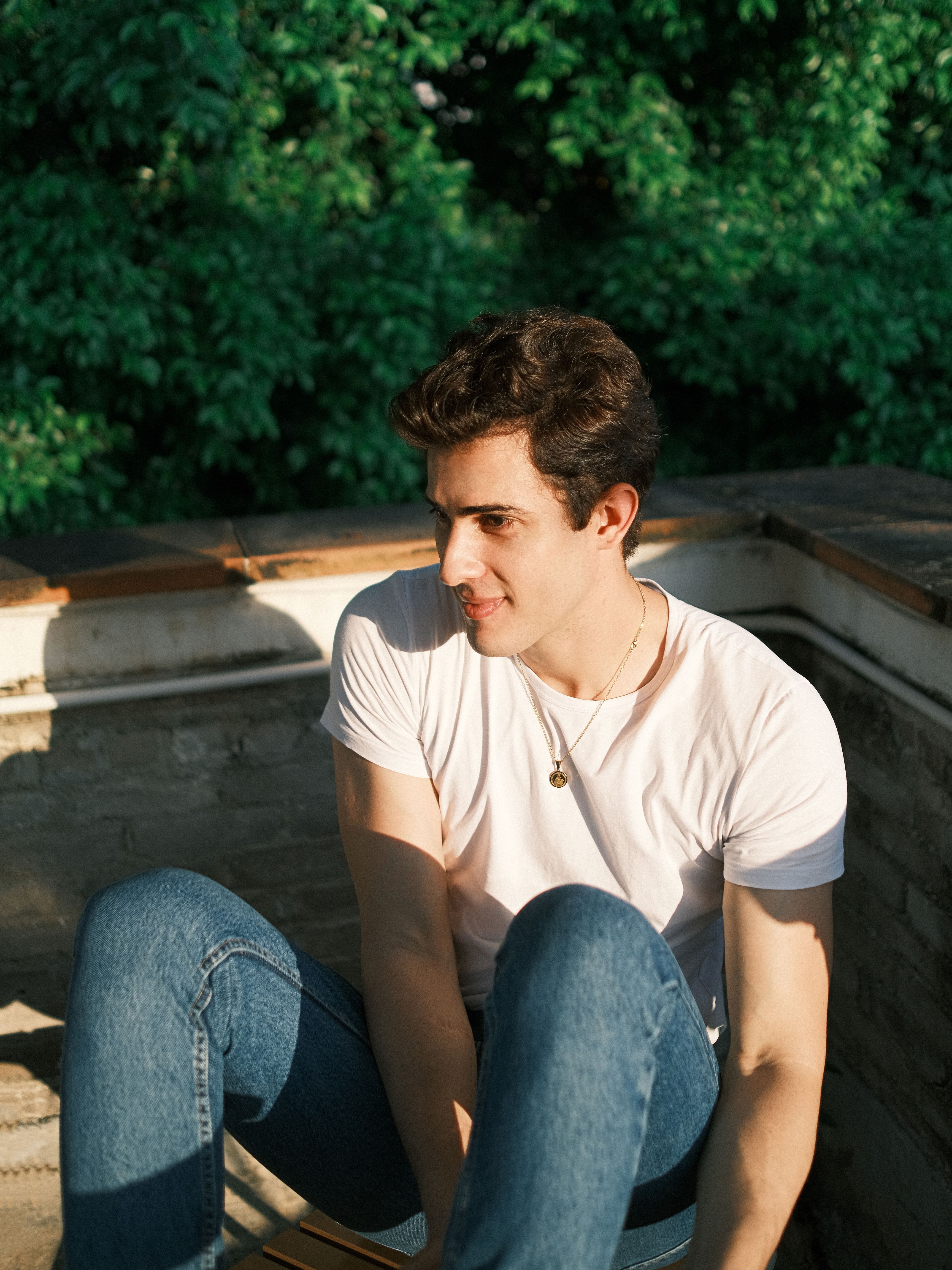Young man in a white t-shirt and jeans sitting outside, enjoying the sunlight.