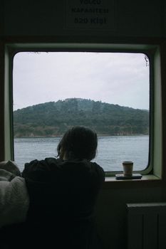 A person gazes out a ferry window at a serene water landscape, evoking a sense of wanderlust.