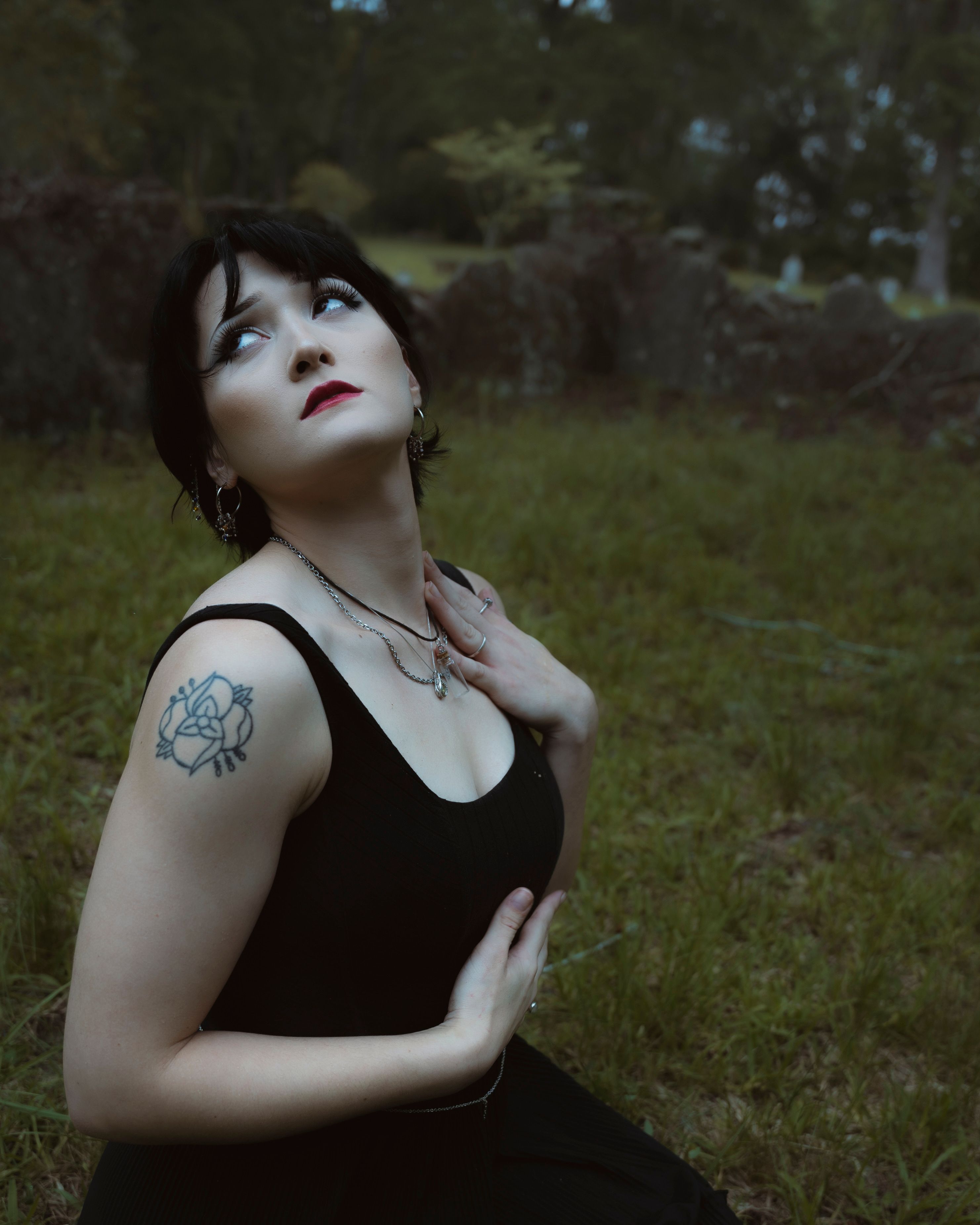 A Gothic-themed portrait of a woman in a black dress with tattoos in a forest setting.