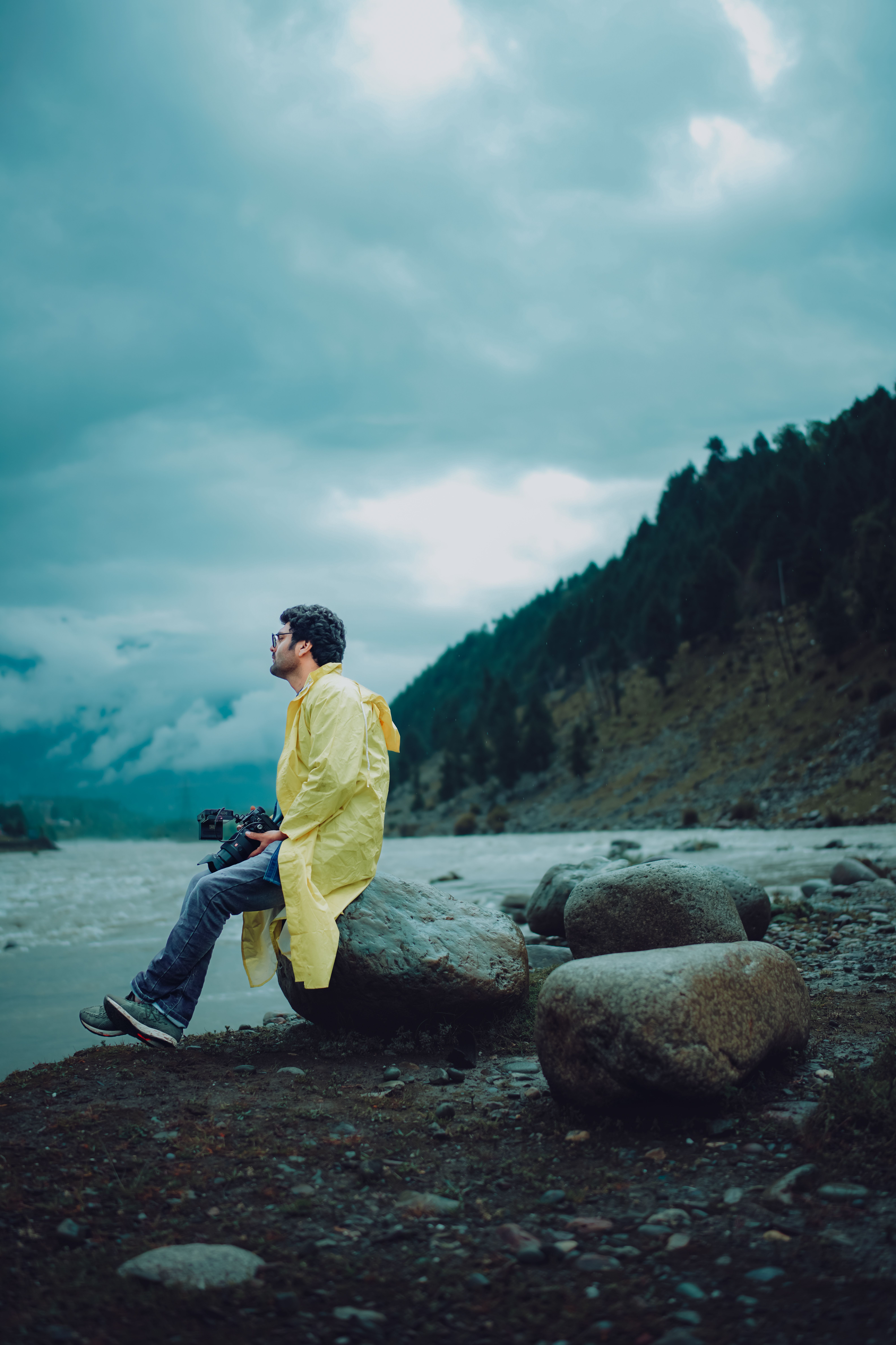 A man in a yellow raincoat sits on a rock by the sea with a camera, under dramatic clouds.