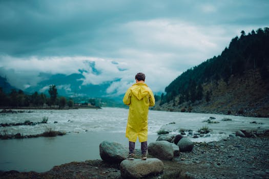 A person stands beside a rushing river with scenic mountains, shrouded in mist.