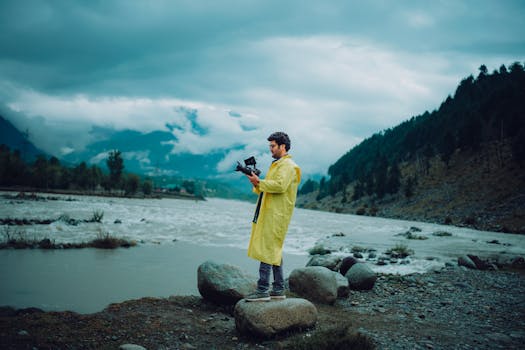 A man in a yellow raincoat stands by a river holding a camera, surrounded by scenic nature.